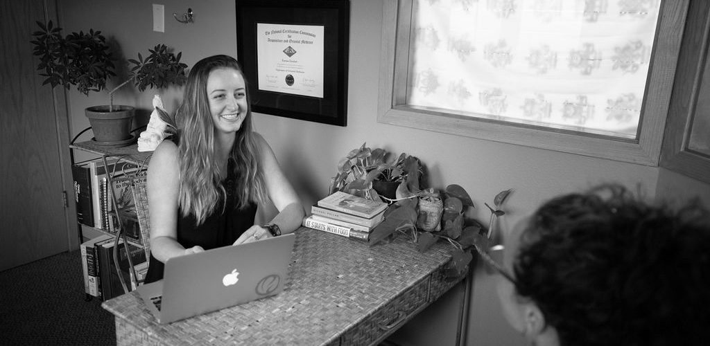 A woman smiles while working at a desk with a dog nearby.